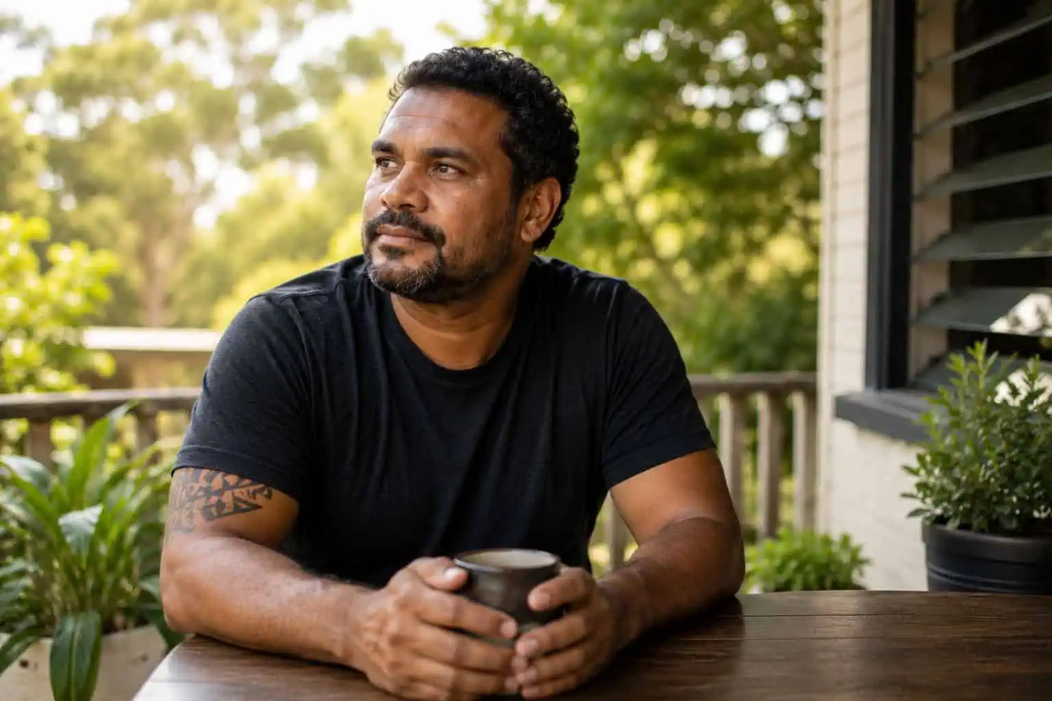 Man sitting thoughtfully on an outdoor deck with a cup of coffee, representing the 715 health check for Aboriginal and Torres Strait Islander people.