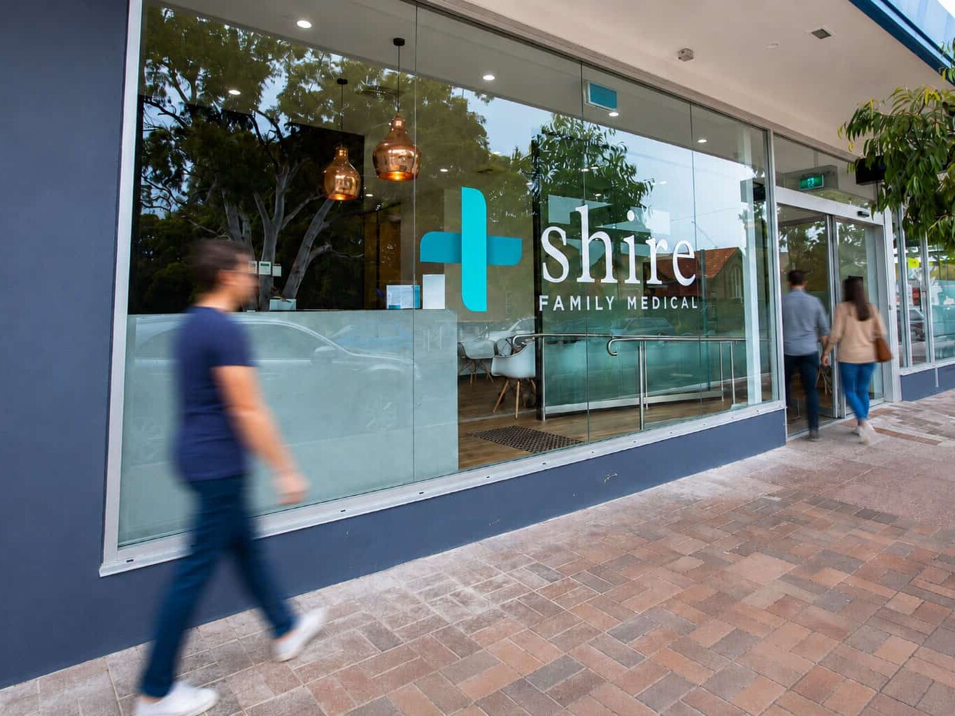 Street view of the Shire Family Medical clinic storefront in Sutherland, showing the navy and teal branded shopfront, glass entrance, and waiting area, with passersby on the footpath.
