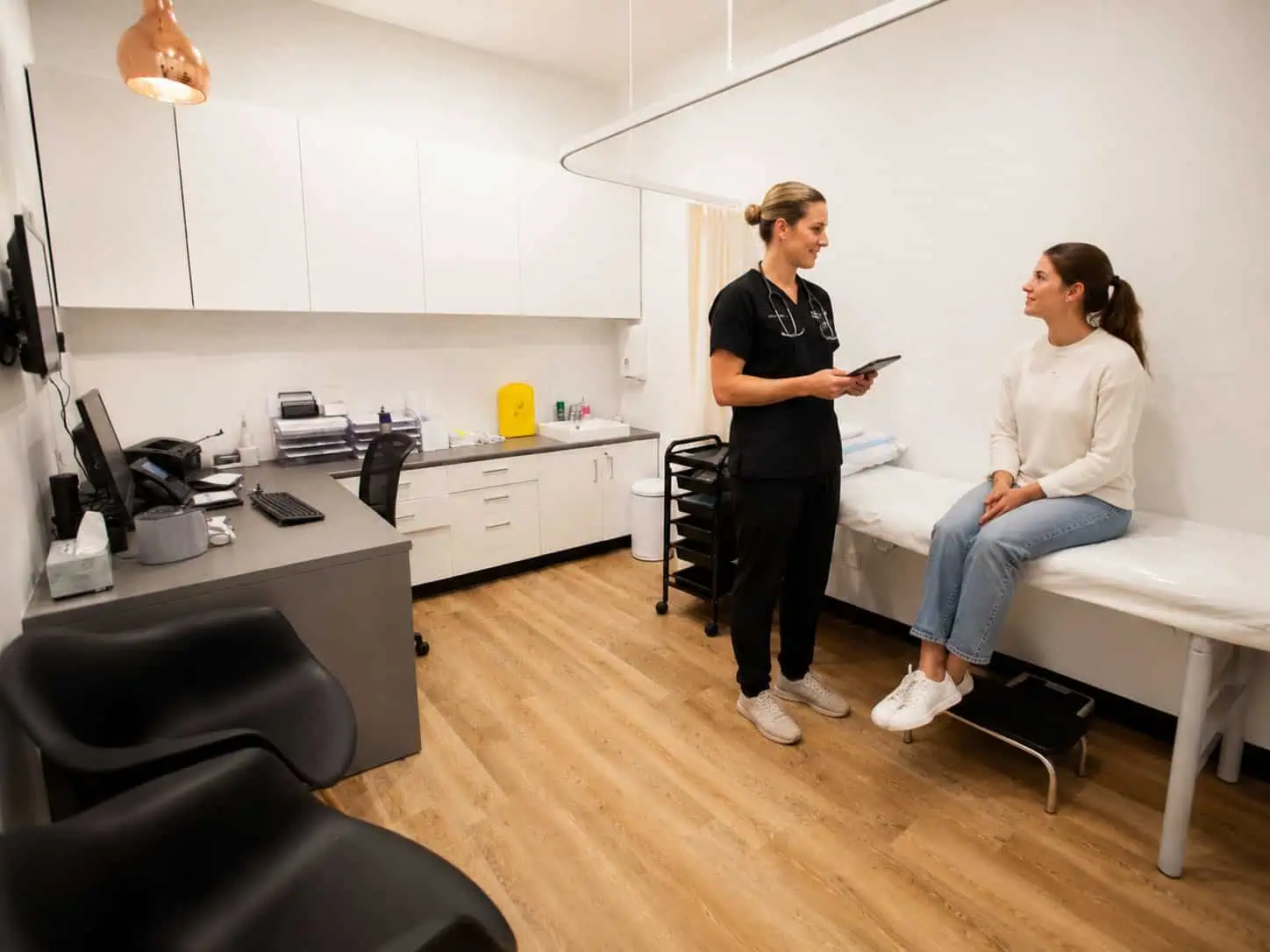 A clinician speaks with a patient seated on the examination table in a bright, modern consulting room at Shire Family Medical.