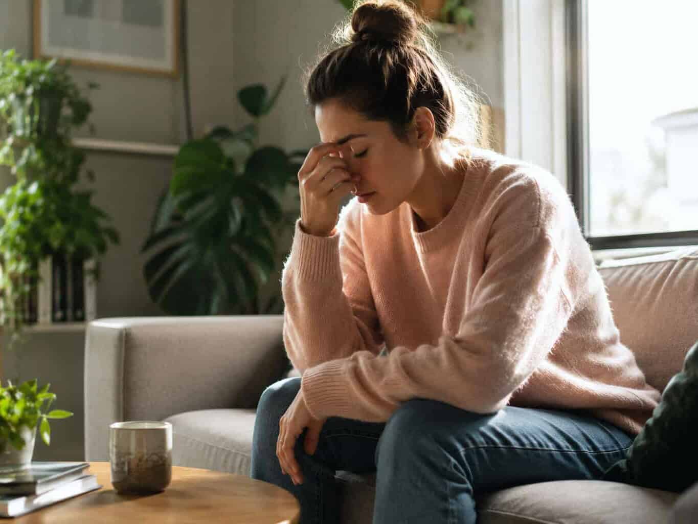 Woman sitting on the couch pinching the bridge of her nose, looking exhausted and stressed, illustrating ongoing fatigue that may need a GP check.